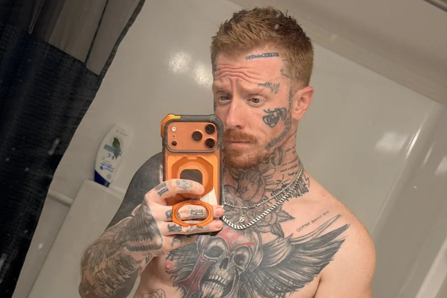 A tattooed man of about 35 takes a confident bathroom mirror selfie, shirtless