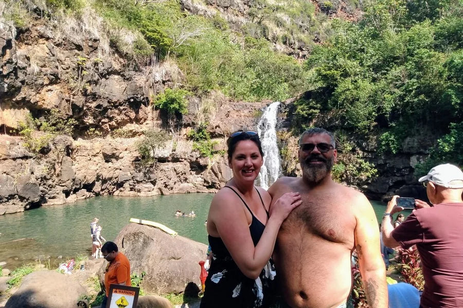 A couple of about 50 smile in front of a waterfall pool, his arm round her