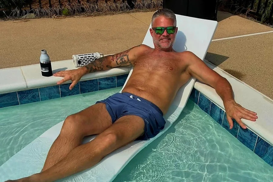 A grey-haired man of about 55 in sunglasses relaxes on a pool lounger with a drink beside him, smiling at the camera
