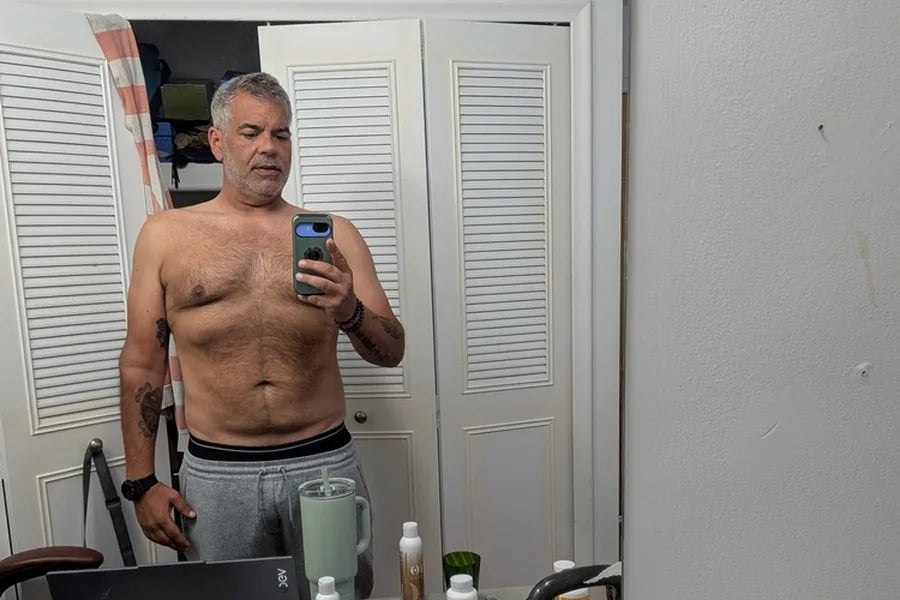 A grey-haired man of about 55 takes a mirror selfie in his home office, supplement bottles on the desk behind him