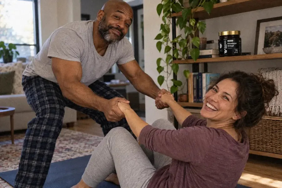 A bald man of about 49 pulls his wife up from a yoga mat by both hands, both laughing, the ShilaSource jar on the shelf behind them